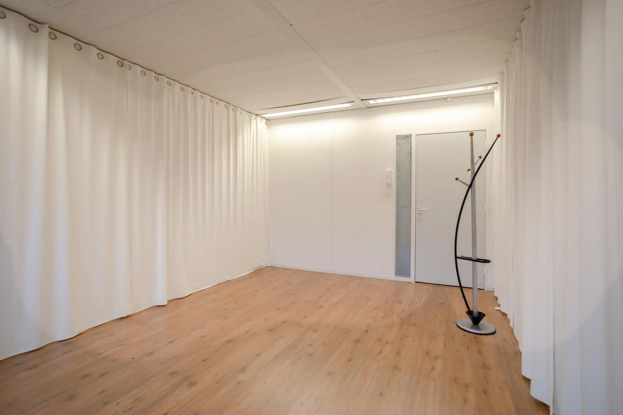 Lichte, lege kamer met houten vloer, witte gordijnen en een moderne kapstok bij een gesloten deur.